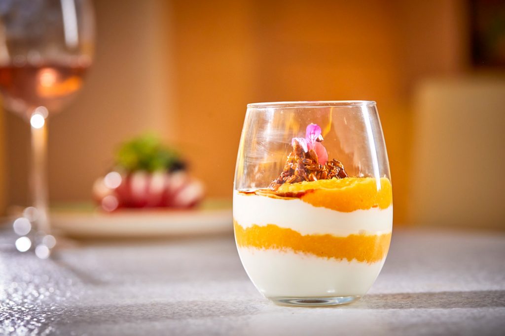 Un primer plano de un postre en una copa sobre una mesa con una copa de vino en el fondo.